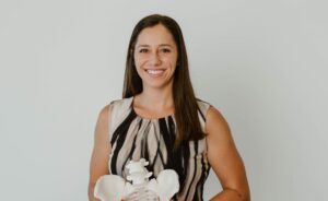Woman with long dark hair smiling while holding an anatomical pelvic model against a light gray background