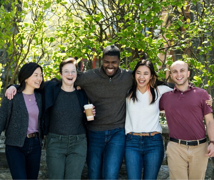 Five students stand arm in arm laughing together outdoors on a sunny campus with green foliage behind them
