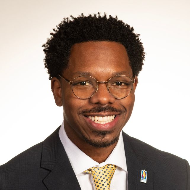 Ravone Cornish smiling in a professional headshot wearing a dark suit and yellow tie with a PCOM lapel pin