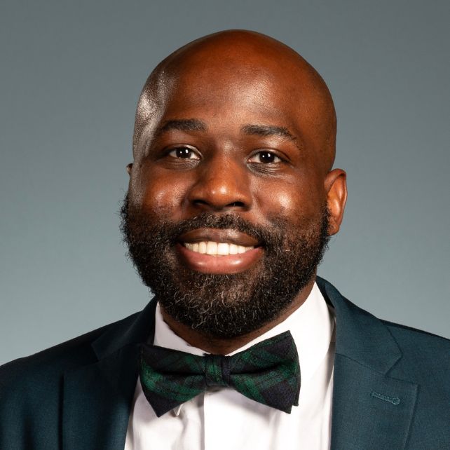 Shavar Simmons smiling in a professional headshot wearing a dark suit and green plaid bow tie against a gray background