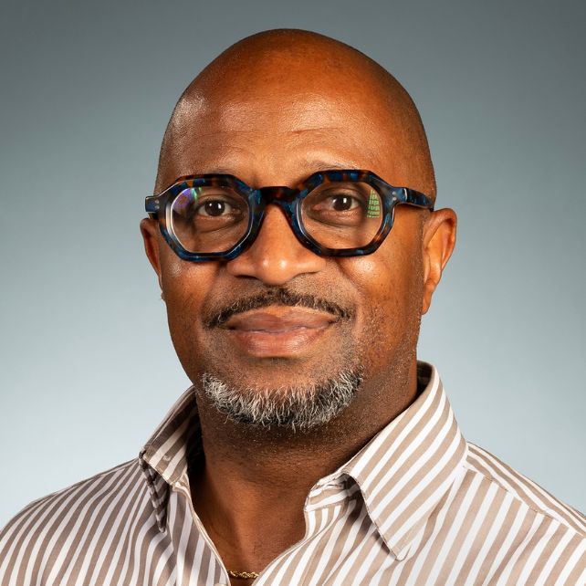 Victor Sawyer smiling in a professional headshot wearing a striped shirt and blue tortoiseshell glasses against a gray background