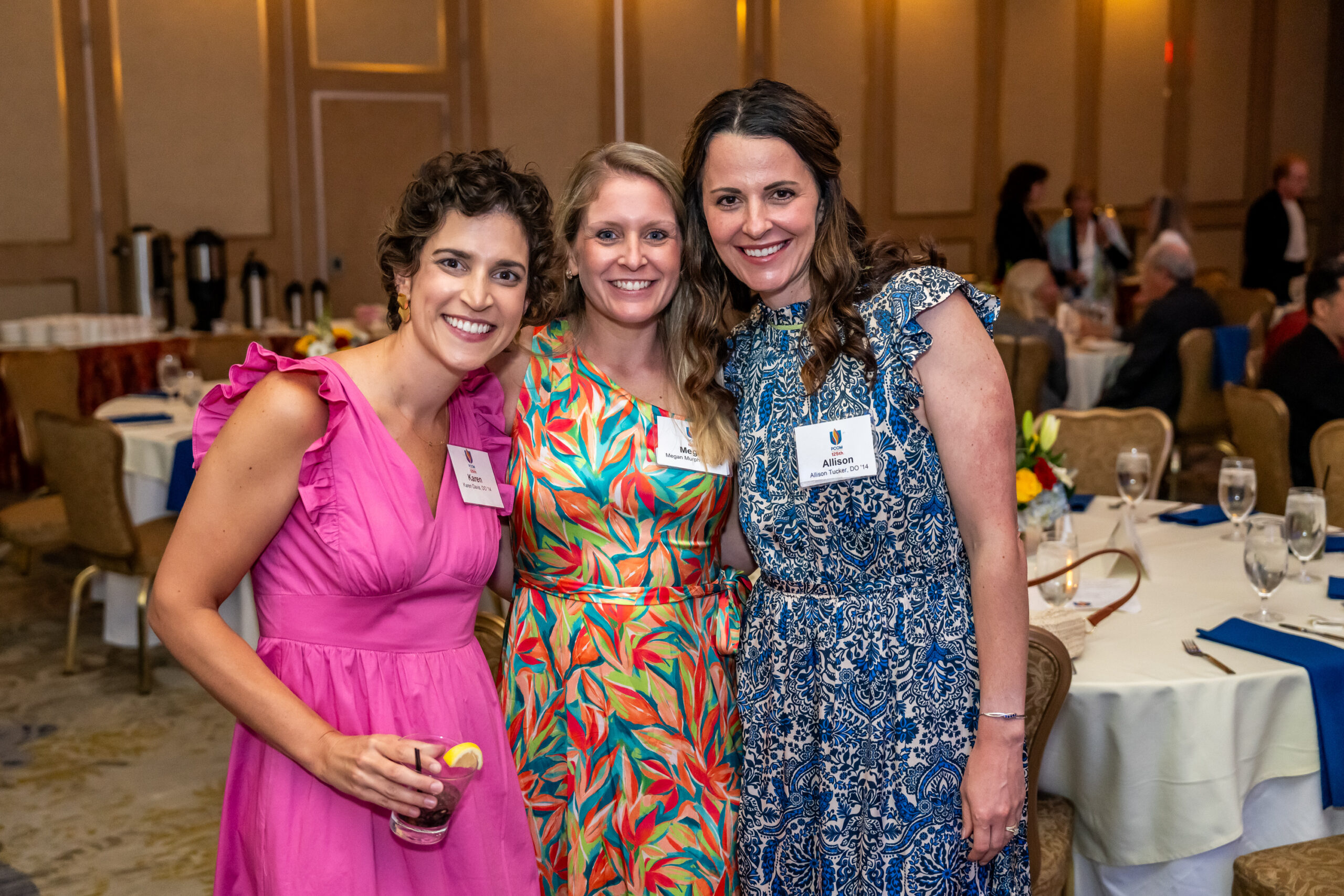 three women dressed up and wearing name badges smile with a ballroom in the background.