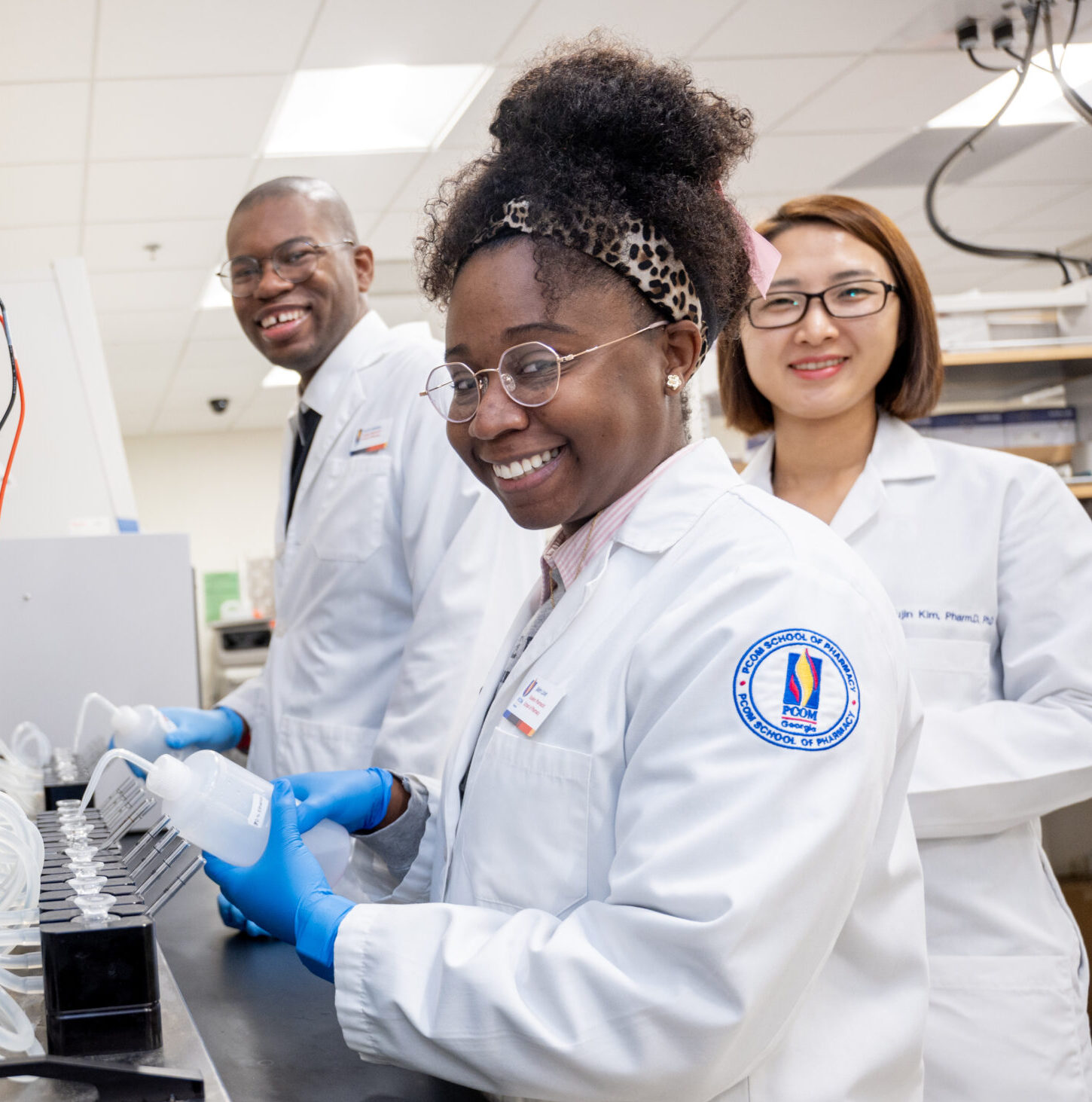 A female pharmacy student is centered with two other students in the background as she works with an instrument in the pharmacy lab.