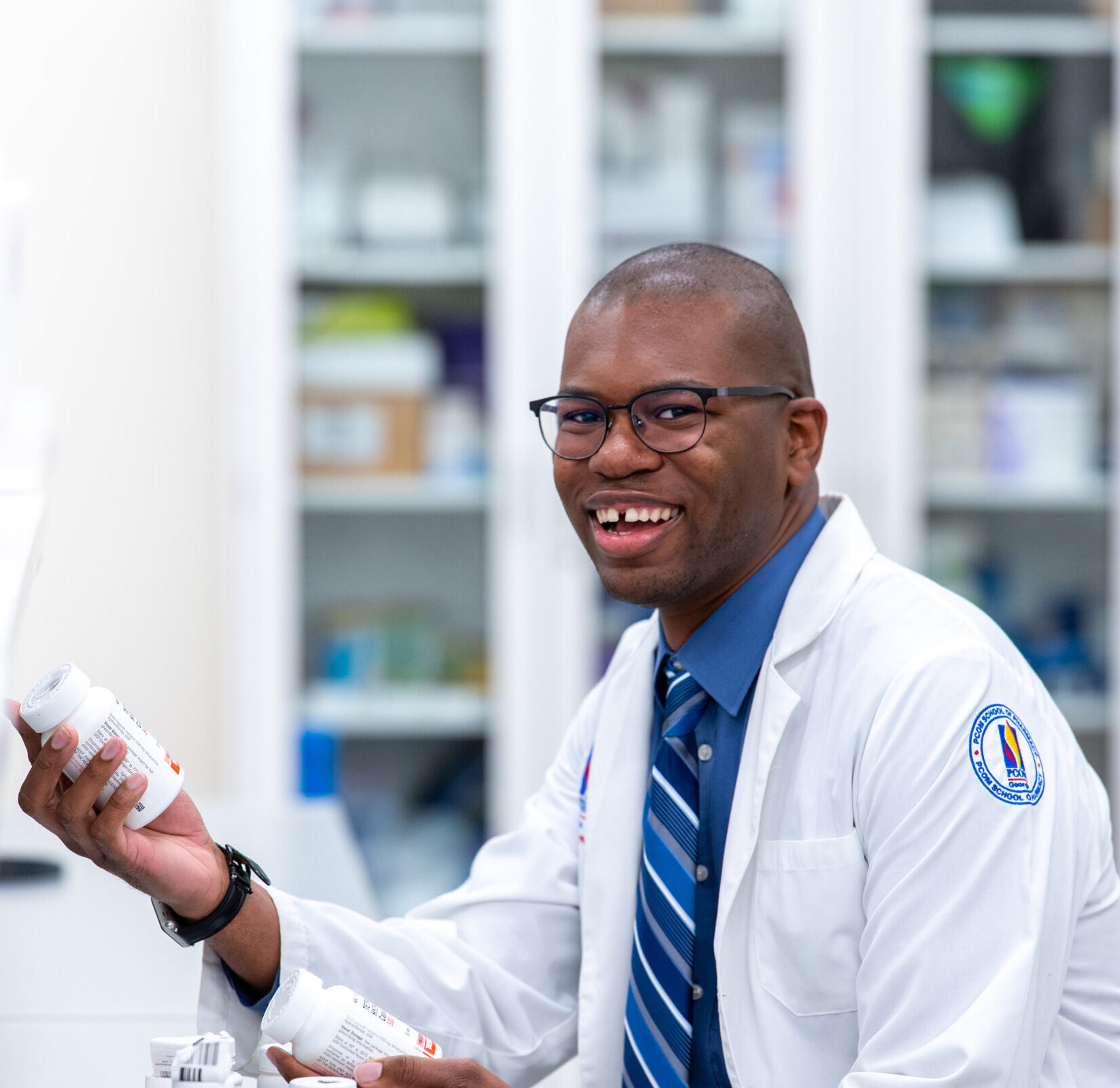 A male student working in the pharmacy lab with prescription bottles smiles while sitting and wearing white coat.