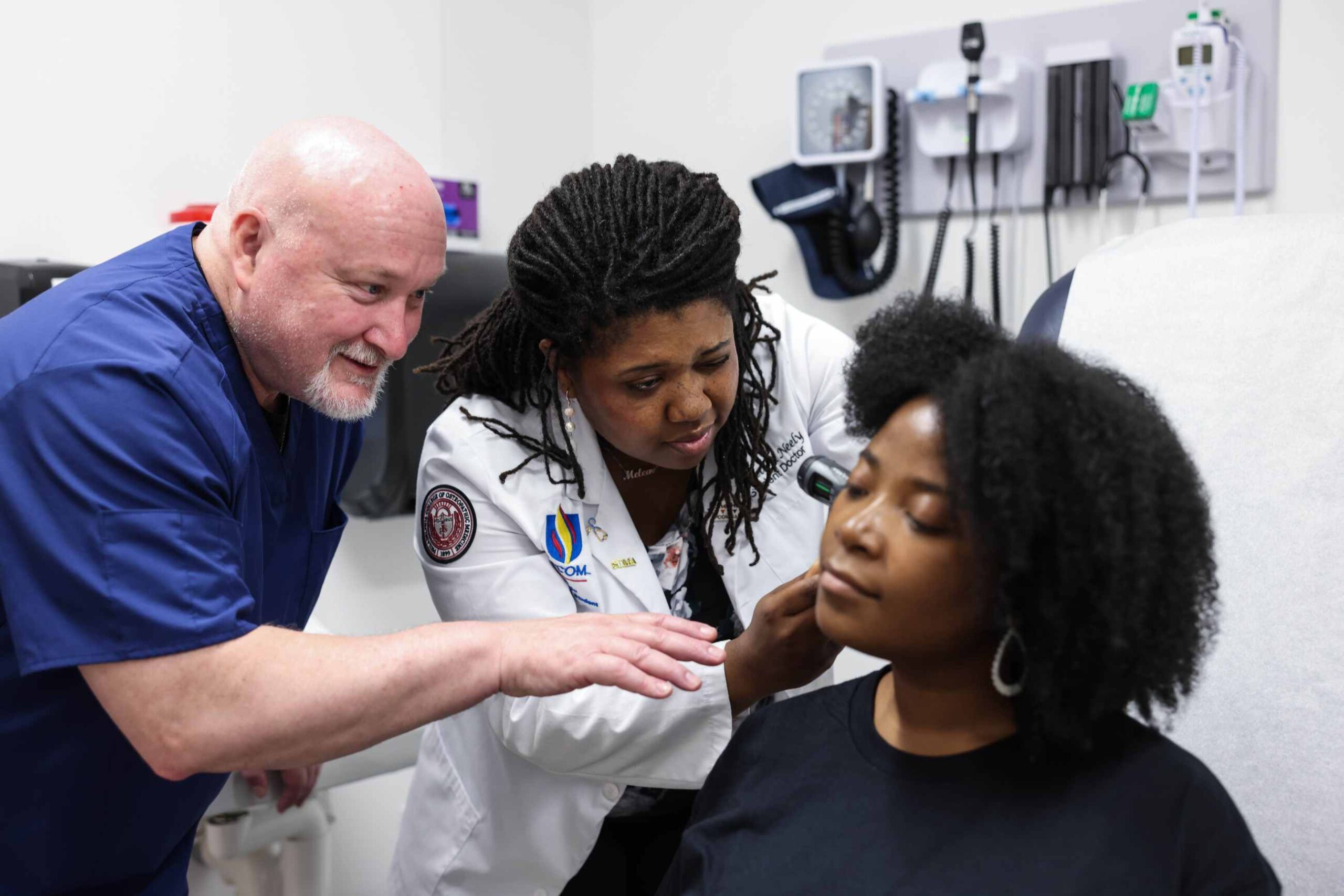 A faculty member guides a medical student in a white coat examining a patient in a clinical setting