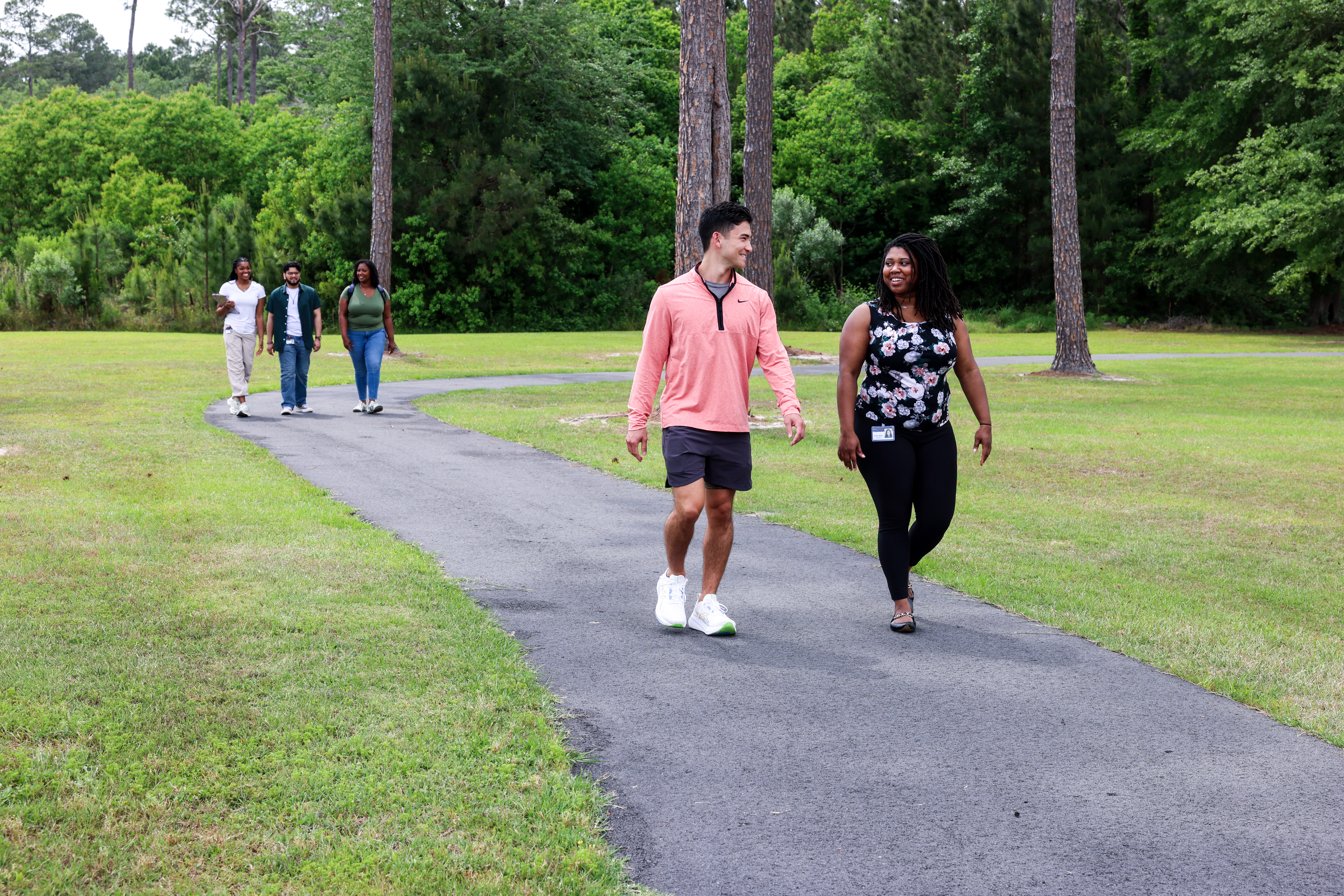 Five students walk along a paved path through a green, tree-lined campus lawn