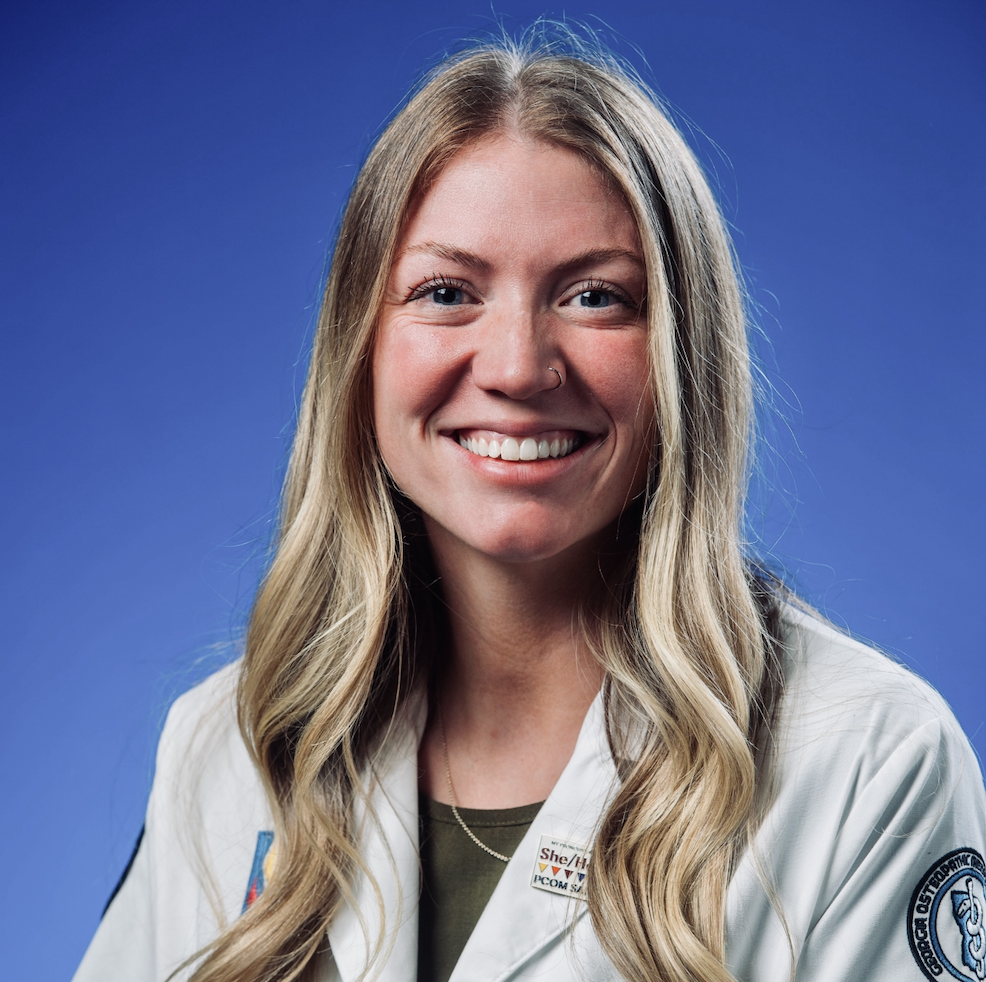 A female student smiles in a white lab coat against a blue background.