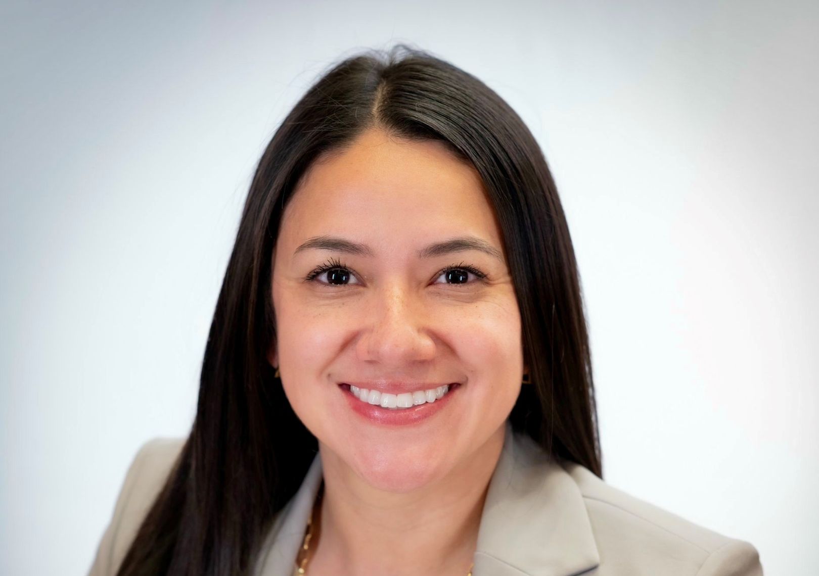 Woman with long dark hair wearing a tan blazer smiling in a professional headshot against a light gray background