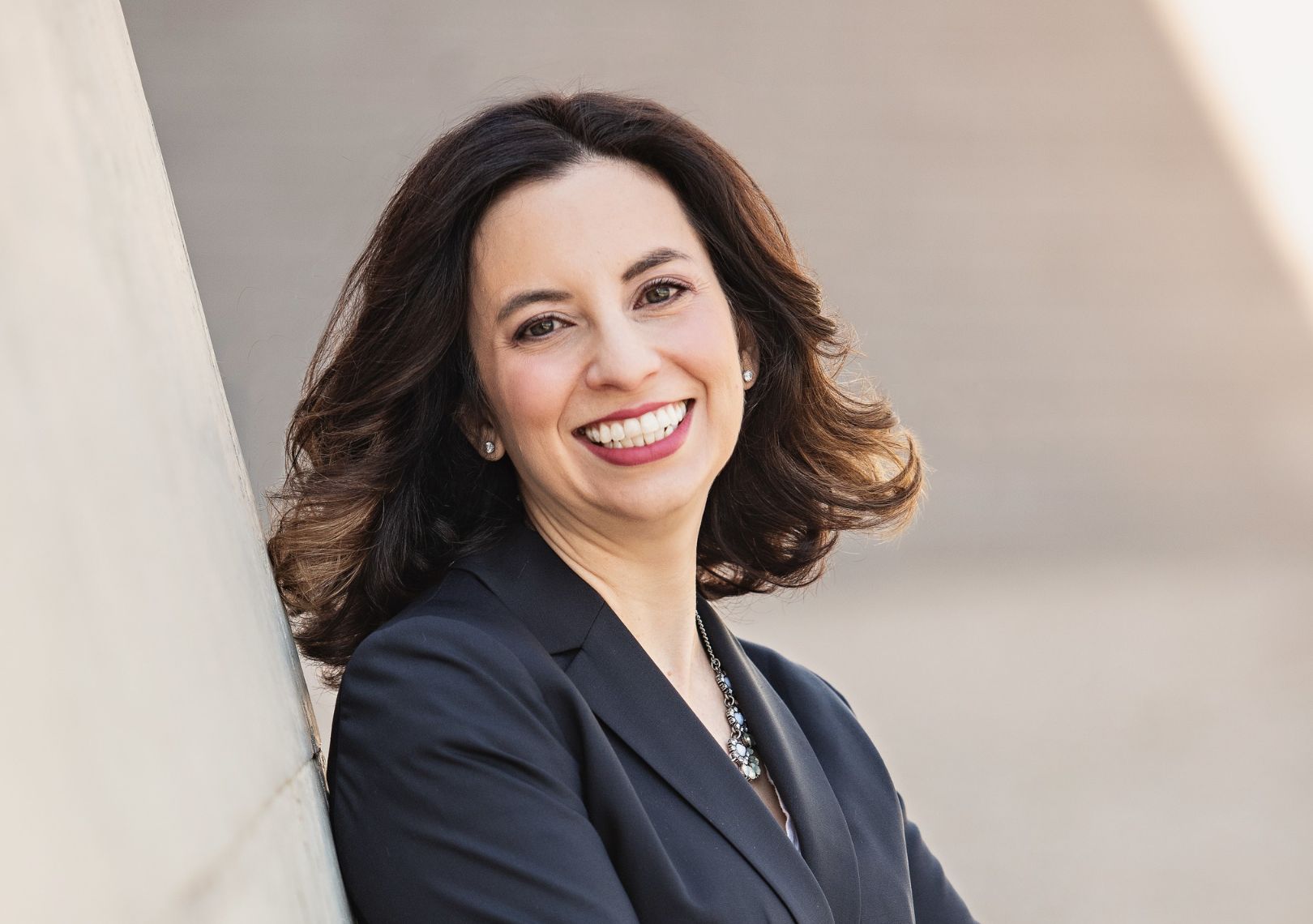 Woman with dark curly hair in a navy blazer and statement necklace smiling in an outdoor professional portrait