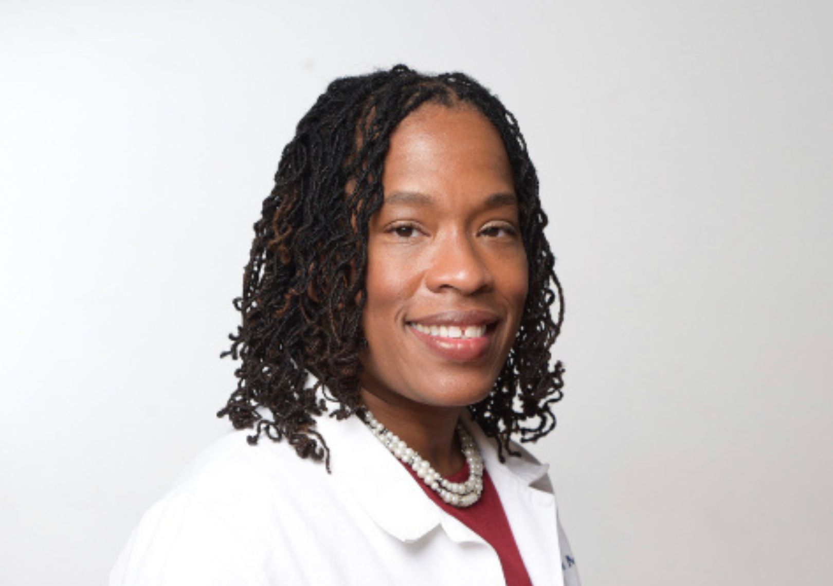 Woman in a white coat and pearl necklace smiling in a professional headshot against a light background