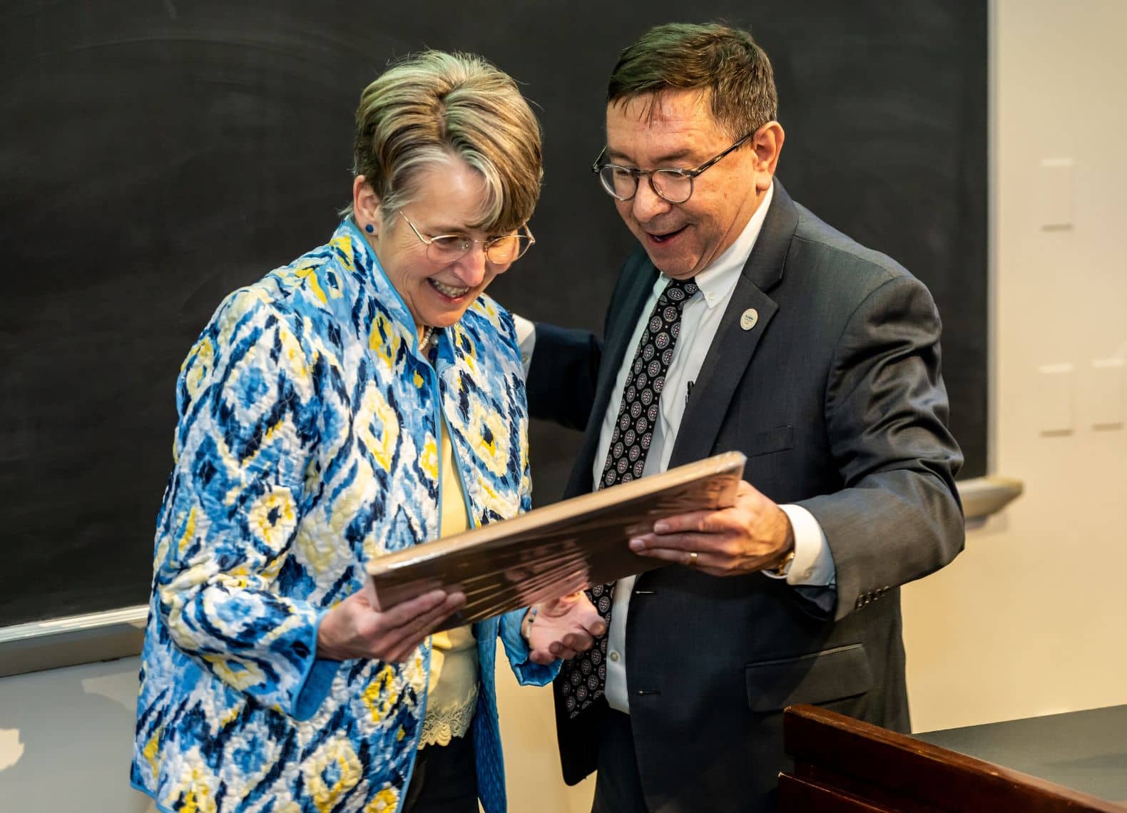 Two people smiling as they look at a plaque together in front of a chalkboard in a classroom