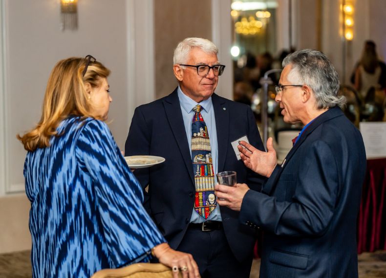 Three people chat over drinks at a formal PCOM alumni reception in a ballroom setting