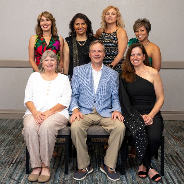 Seven alumni pose for a group photo at a formal PCOM event, three seated in front and four standing behind