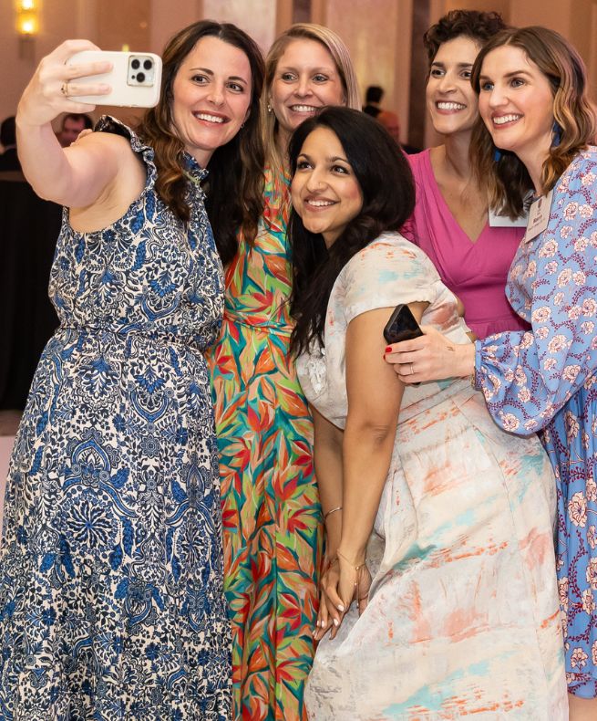 Five women smile and pose for a group selfie at a PCOM alumni event in a ballroom setting