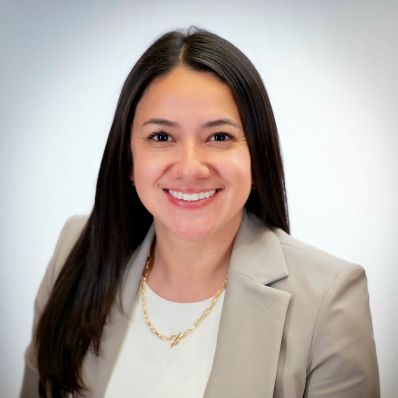 Angie Amado smiling in a professional headshot wearing a beige blazer and gold chain necklace against a light gray background
