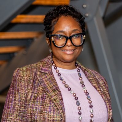 Ashara Calisse-Atchley Cashaw smiling in a professional headshot wearing a tweed blazer, beaded necklace, and glasses