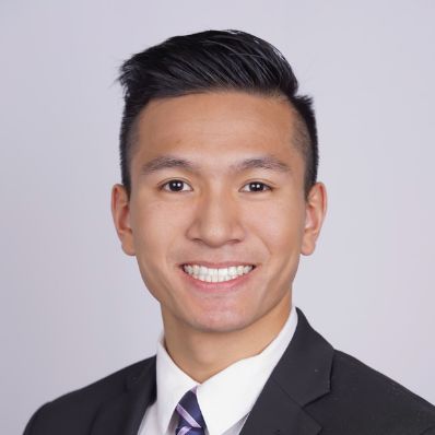 Brandon Dang smiling in a professional headshot wearing a dark suit and striped tie against a light gray background