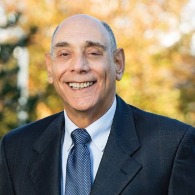 Man in a dark suit and blue polka-dot tie smiling outdoors with autumn foliage in the background