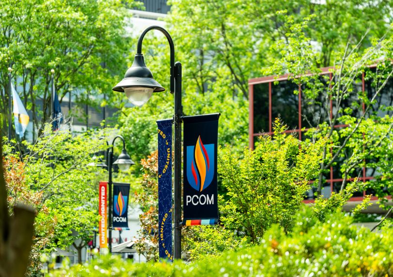 PCOM branded banners on lampposts surrounded by lush green trees on the campus grounds