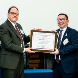 Two men in suits smile while presenting a framed PCOM certificate of honor at an awards ceremony