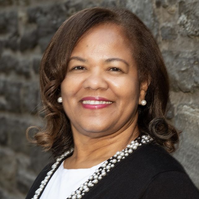 Chandra Tyler smiling in a professional headshot wearing a black blazer and pearl necklace against a stone wall backdrop