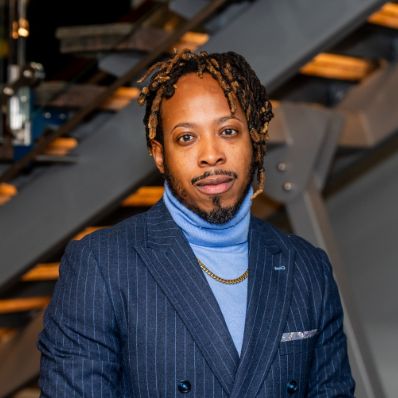 Christopher Brown in a professional headshot wearing a navy pinstripe blazer and light blue turtleneck with a gold chain