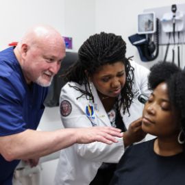A faculty member guides a medical student in a white coat examining a patient in a clinical setting