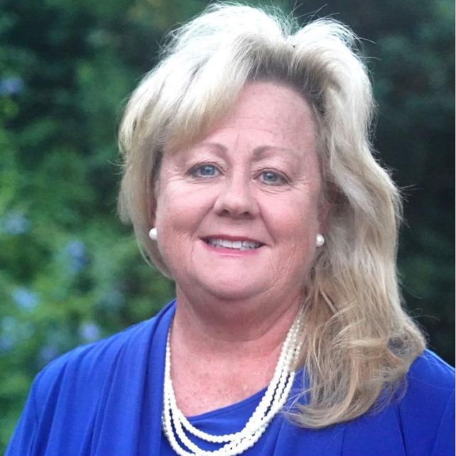 Deidre Mercer Martin smiling in a headshot wearing a blue top and pearl necklace with green foliage in the background