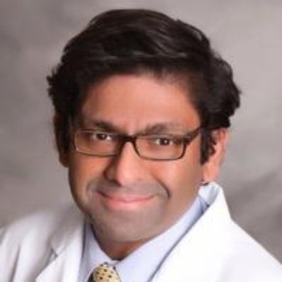 Dipan Desai smiling in a professional headshot wearing a white coat, yellow tie, and glasses against a gray background