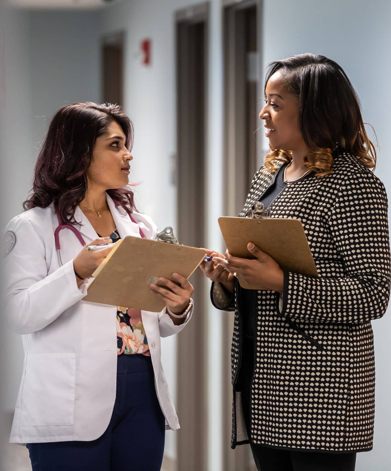 A doctor in a white coat with a stethoscope and a colleague holding clipboards discuss in a hospital hallway