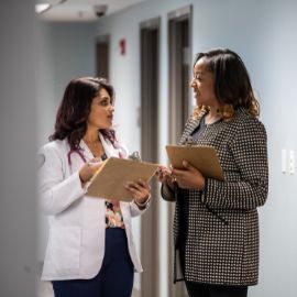 A woman in a white coat and a colleague holding clipboards discuss in a hospital hallway