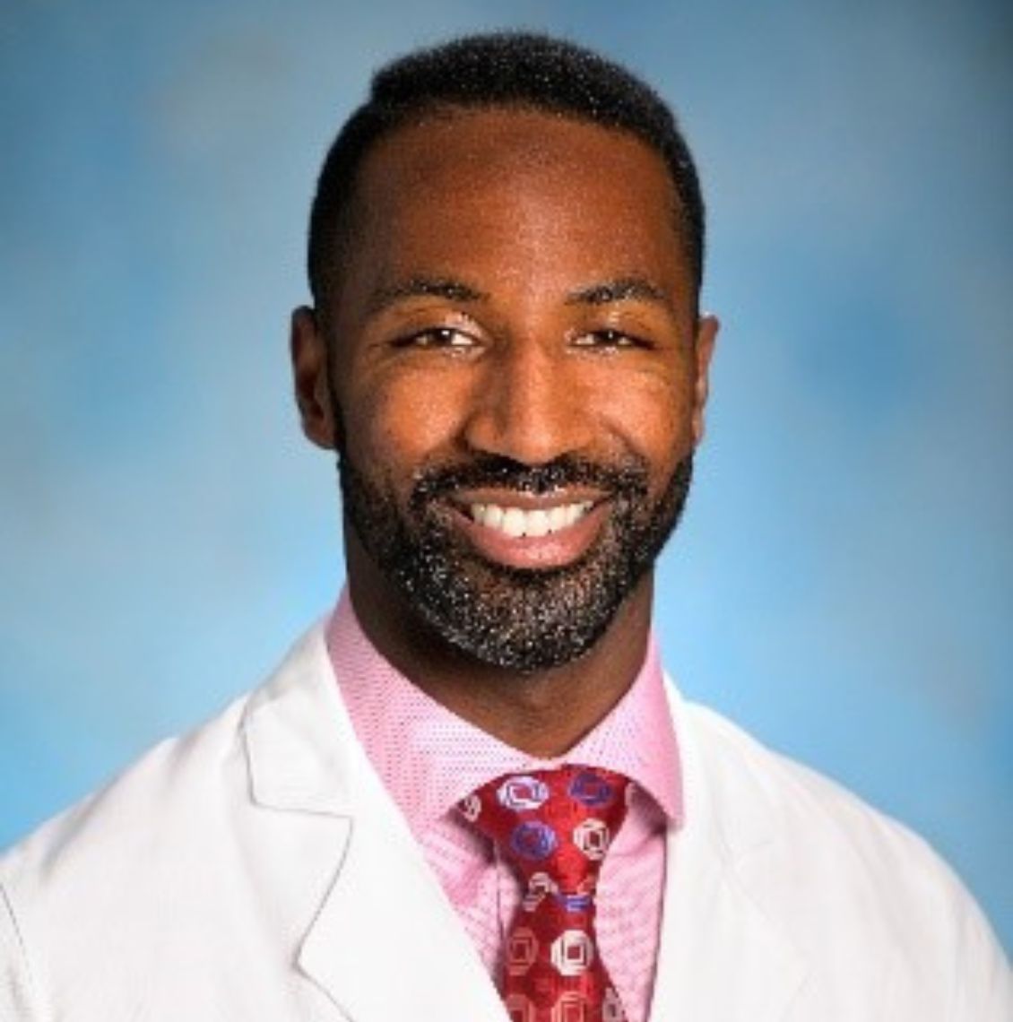 Man in a white coat, pink shirt, and red patterned tie smiling in a professional headshot against a blue background