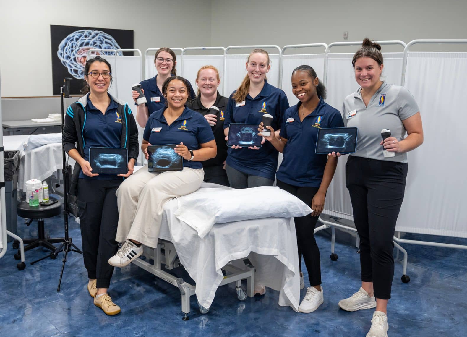 Eight PCOM students in navy polos pose with ultrasound tablets and probes around an exam table in a simulation lab