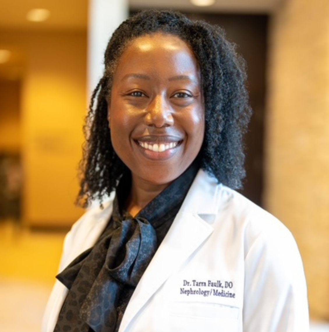 Dr. Tarra Faulk, DO, smiling in a white coat embroidered with Nephrology/Medicine in a modern building lobby