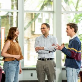 A faculty member and two students chat in a bright glass-walled campus atrium