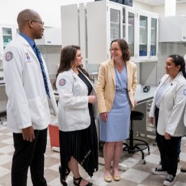 Faculty and students in white coats converse with a colleague in a campus laboratory setting