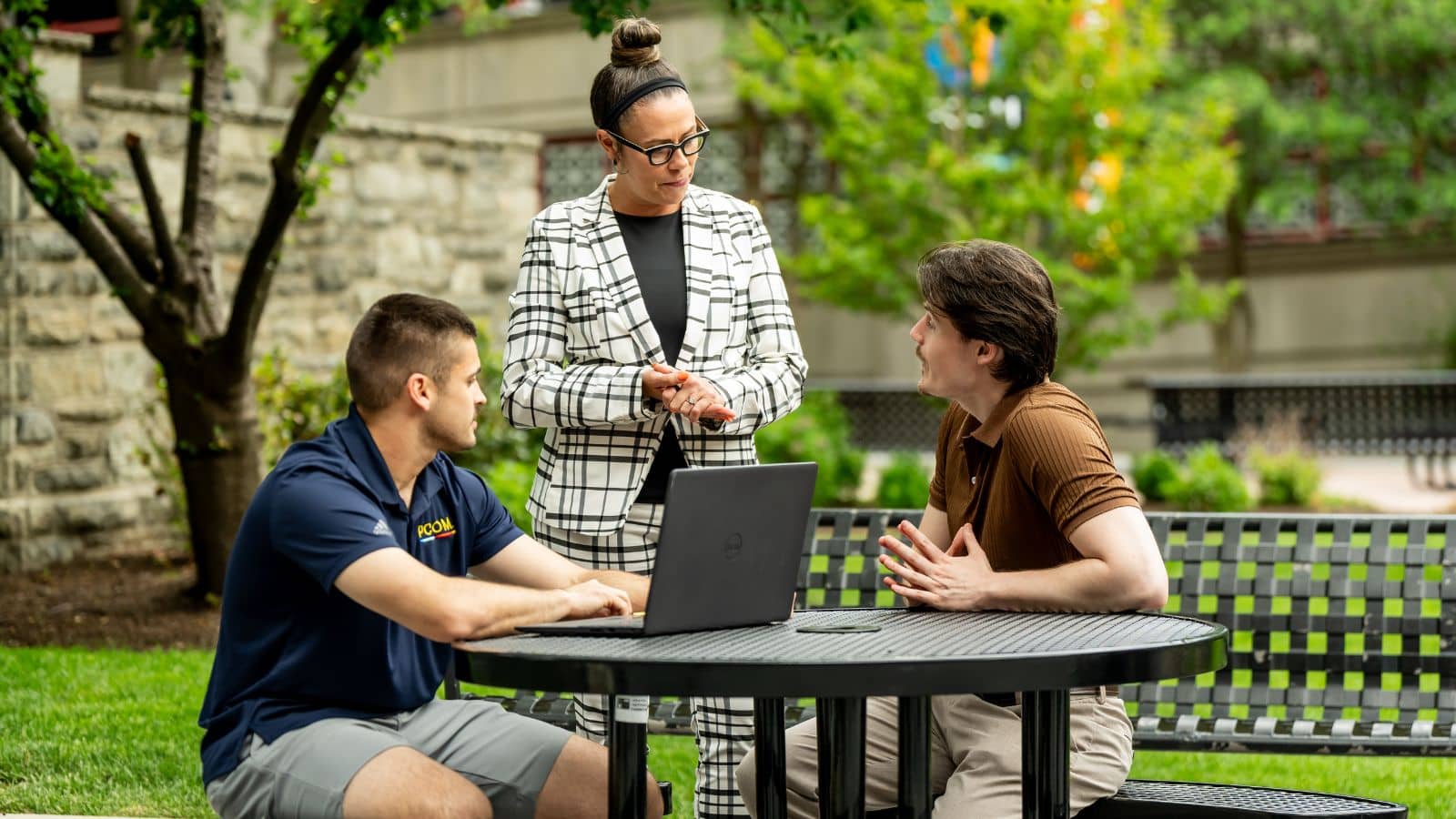 A faculty member in a plaid blazer speaks with two students at an outdoor campus table with a laptop