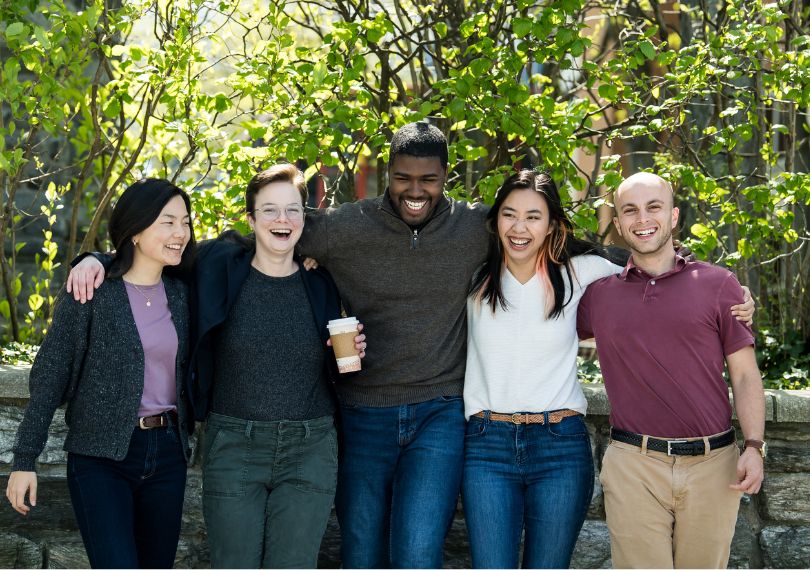 Five students stand arm in arm laughing together outdoors on a sunny campus with green foliage behind them
