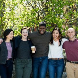 Five students stand arm in arm laughing together outdoors on a sunny campus with green foliage behind them