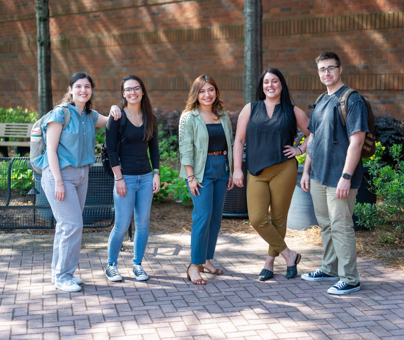 Five students pose together on a sunny brick courtyard with a campus building and garden beds behind them