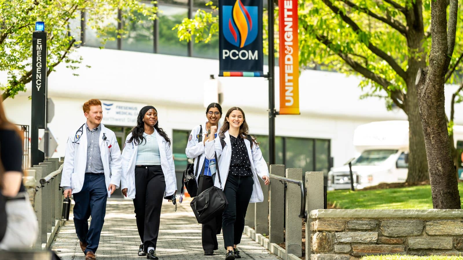 Four medical students in white coats with stethoscopes walk together along a campus path with PCOM banners overhead