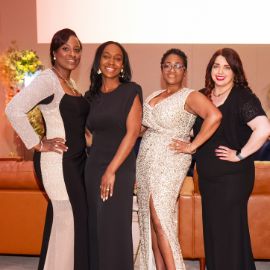 Four women in elegant evening gowns pose together at a formal PCOM gala event