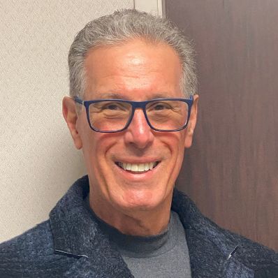 Glenn Freed smiling in a headshot wearing a gray sweater, dark jacket, and blue-framed glasses