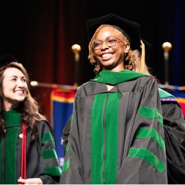 Smiling graduate in black and green doctoral regalia walks across the commencement stage
