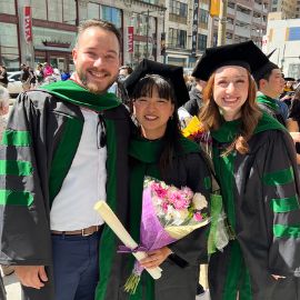 Three people celebrate at a PCOM commencement, two wearing black and green graduation caps and gowns while holding flowers and a diploma