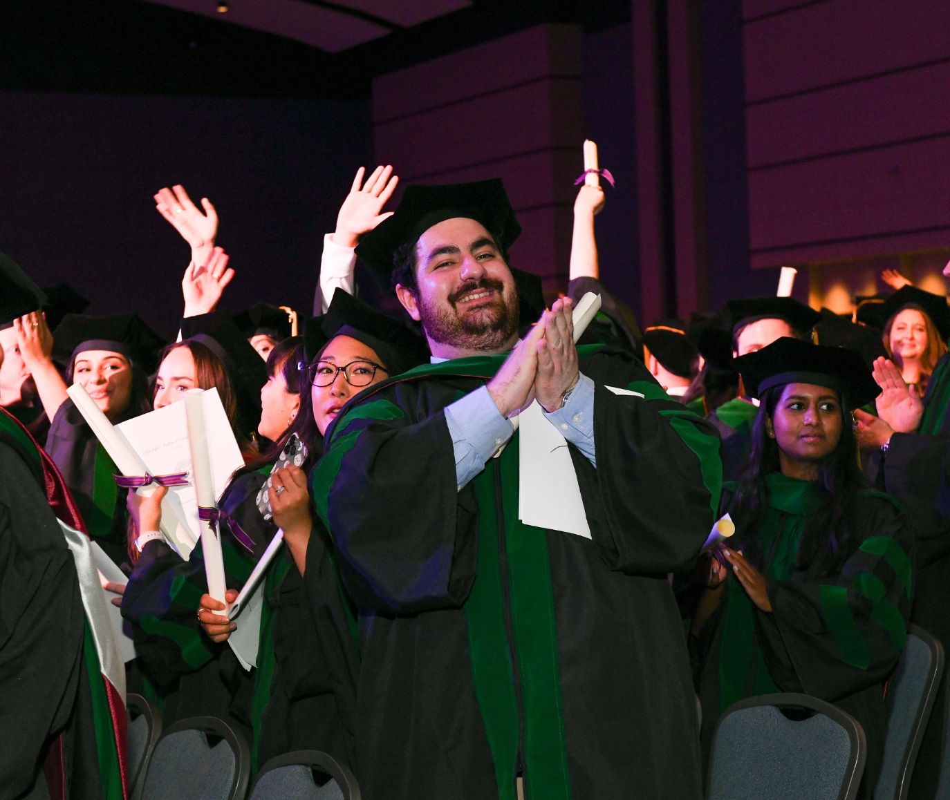 PCOM graduates in black and green caps and gowns clap and wave their diplomas during a commencement ceremony