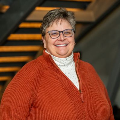 Gretta Gross smiling in a professional headshot wearing an orange quarter-zip sweater and glasses in a modern building lobby