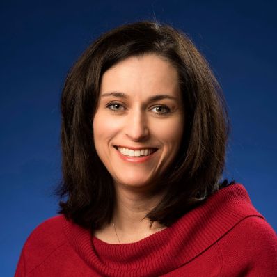 Woman with dark brown hair wearing a red sweater smiling in a professional headshot against a blue background
