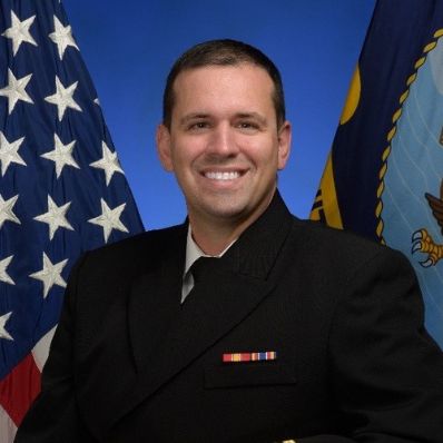 Man in a Navy dress uniform smiling with arms crossed in front of American and military flags