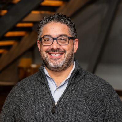 Joshua Baron smiling in a professional headshot wearing a gray cable-knit sweater and glasses in a modern building lobby